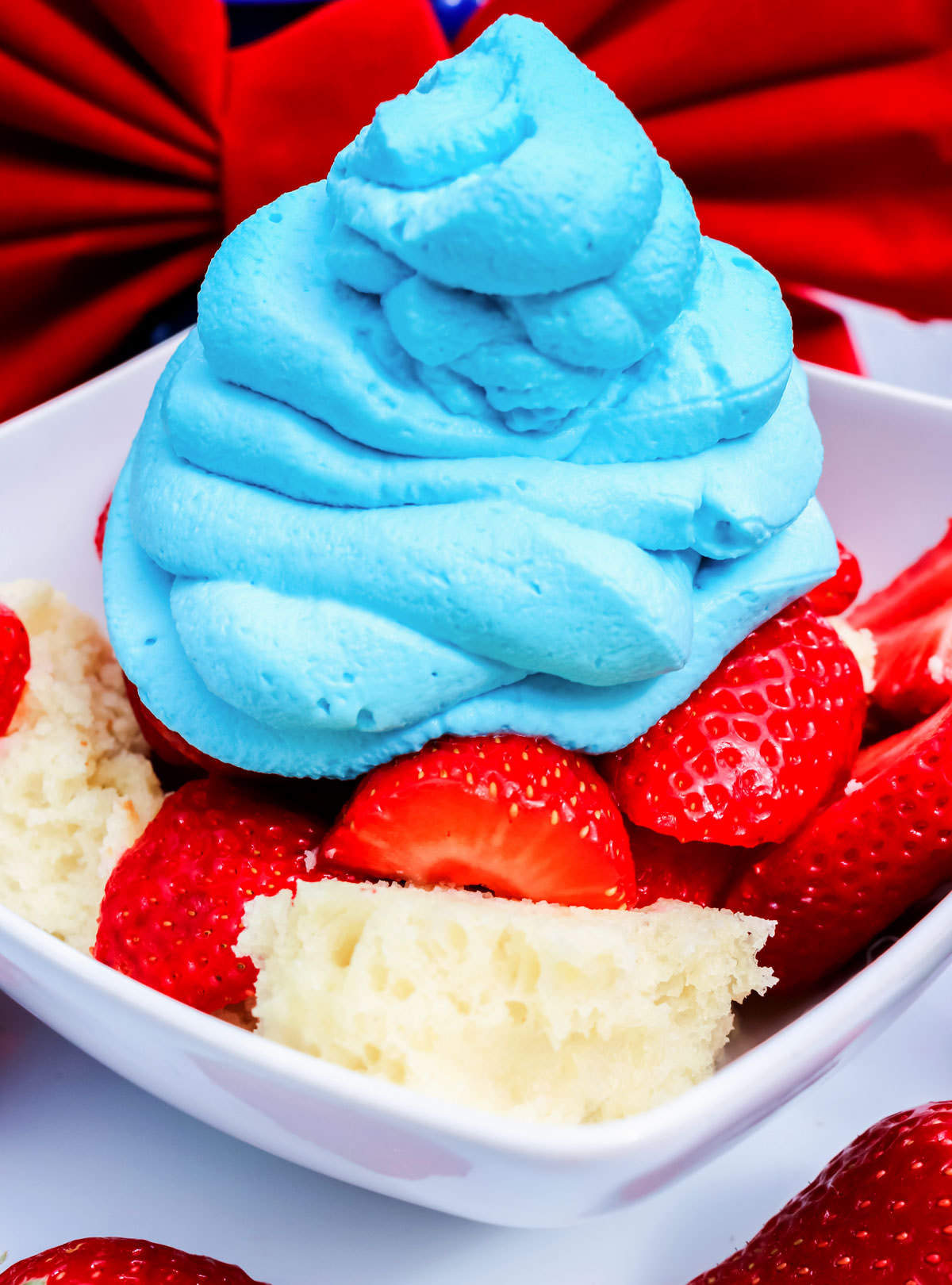 Closeup of a white bowl filled with Red White and Blue Strawberry Shortcake sitting on a white table in front of a big red bow.