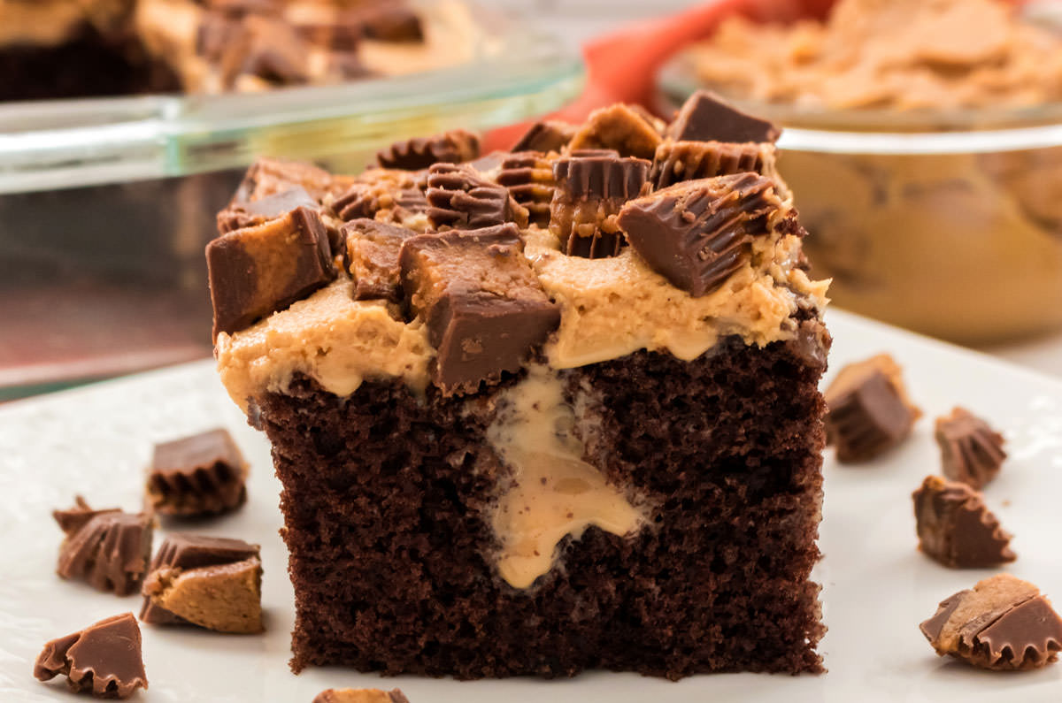 Closeup on a piece of Chocolate Peanut Butter Poke Cake sitting on a white plate in front of the cake pan and a glass bowl filled with Peanut Butter.