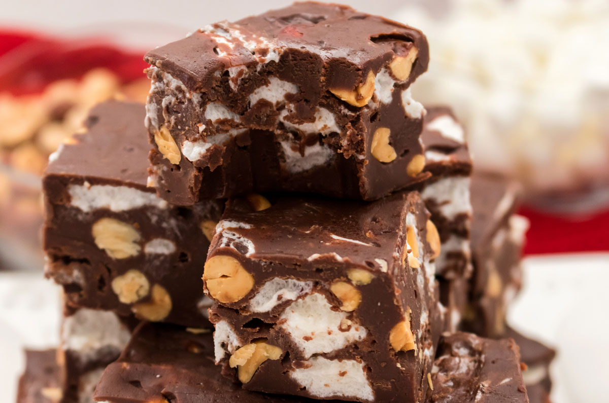 Closeup on a stack of Rocky Road Fudge pieces sitting on a white surface in front of glass bowls filled with peanuts and marshmallows.
