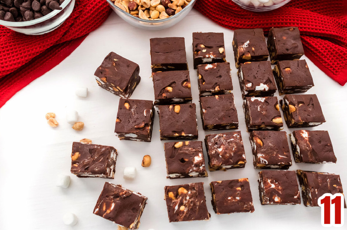 Overhead shot of 24 pieces of Rocky Road Fudge lined up in rows on a white table surrounded by peanuts and marshmallows.