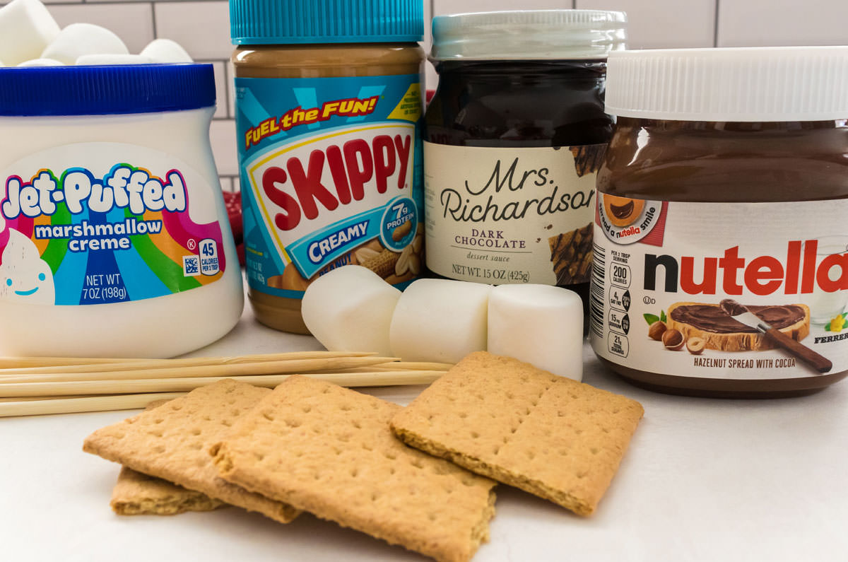 Jars of Marshmallow Creme, Peanut Butter, Chocolate Sauce and Nutella sitting on a white table along with graham crackers and marshmallows.