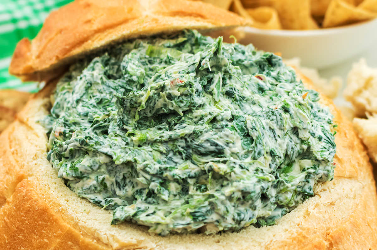 Close up of Creamy Spinach Dip served in a hollowed up Sourdough Round, surrounded by chips and bread pieces.