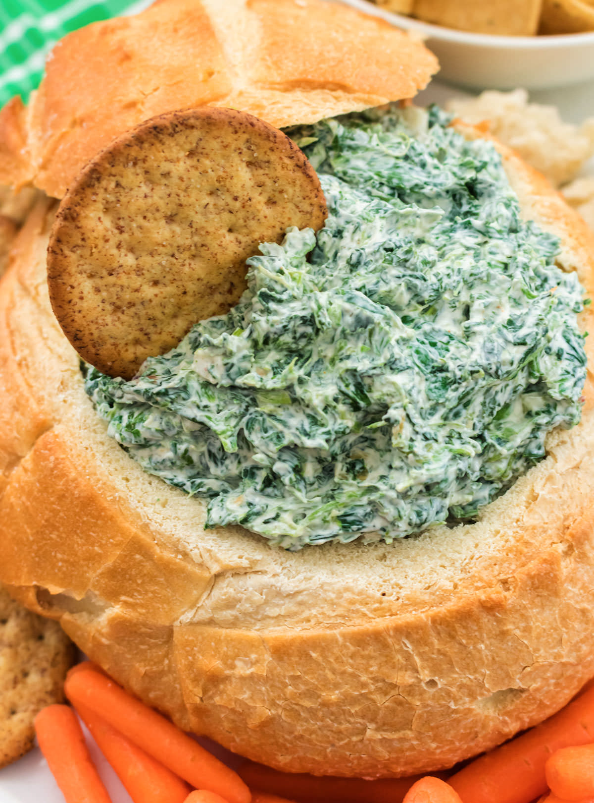 Closeup on a batch of Spinach Dip in a hollowed up loaf of sourdough bread surrounded by carrots, crackers and pieces of bread.