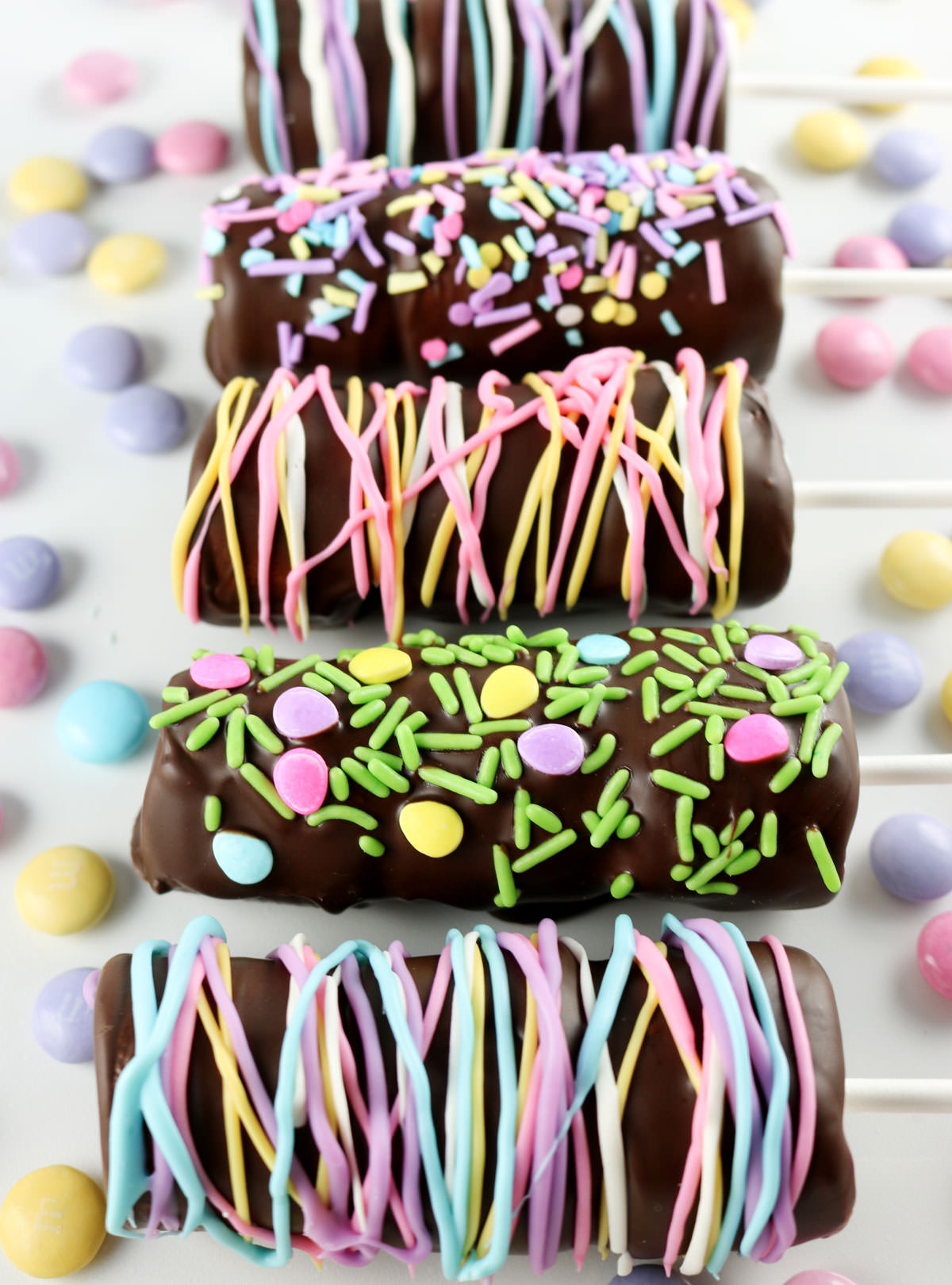 Closeup on six Springtime Marshmallow Pops laying on a white surface surrounded by M&Ms.