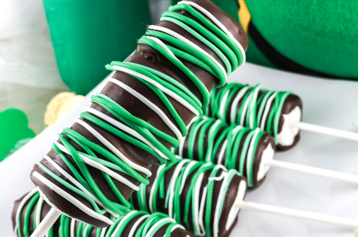 Closeup on a St. Patrick's Day Marshmallow Pop being held over a plate full of other marshmallow pops in front of a leprechaun hat.