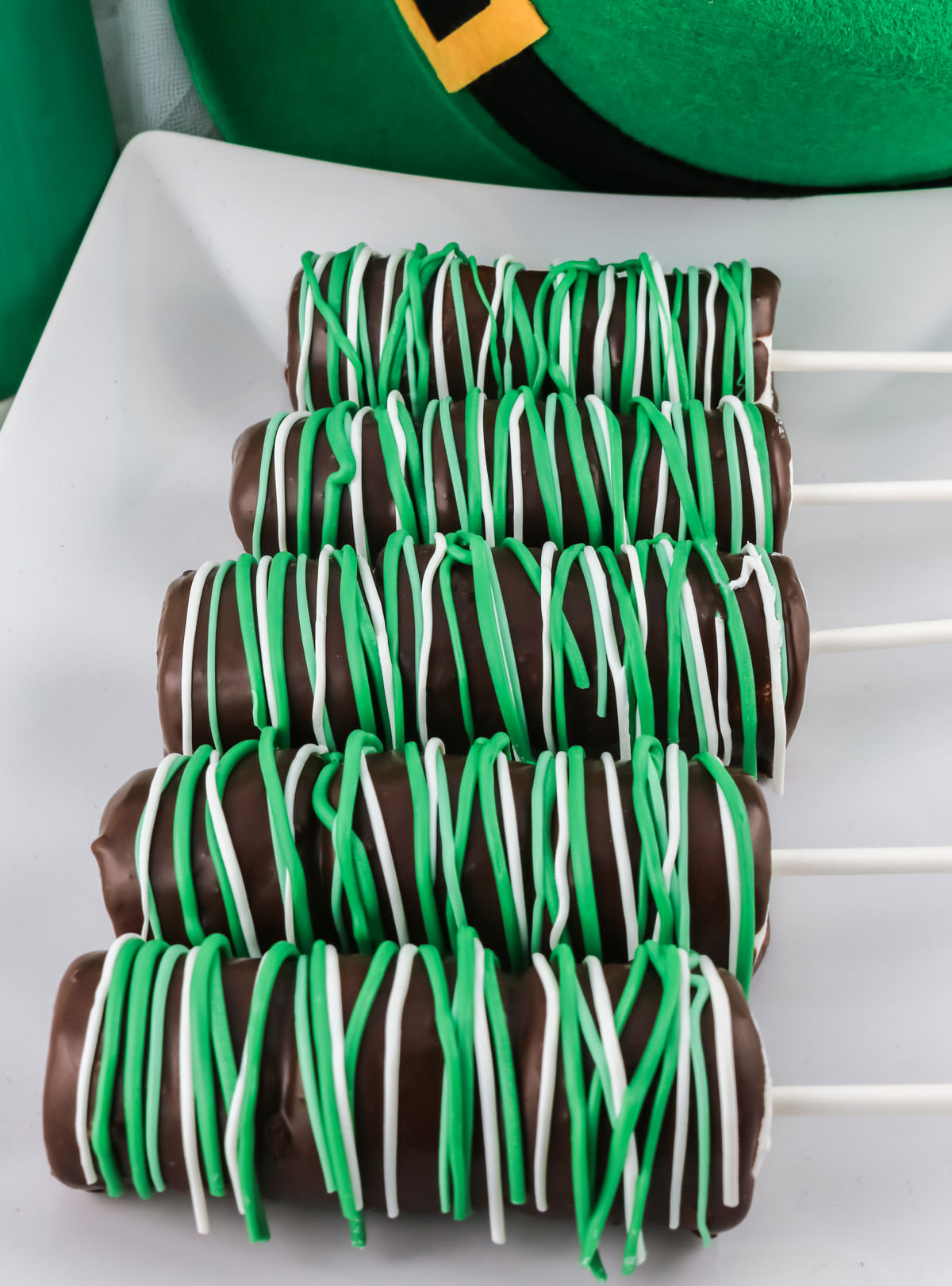 Five St. Patrick's Day Marshmallow Pops laying on a white serving platter in front of a Leprechaun hat.