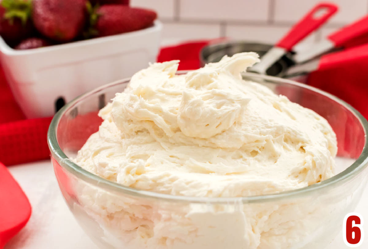 Closeup on a glass bowl filled with Whipped Vanilla Frosting sitting in front of a red towel and a ceramic basket filled with fresh strawberries.