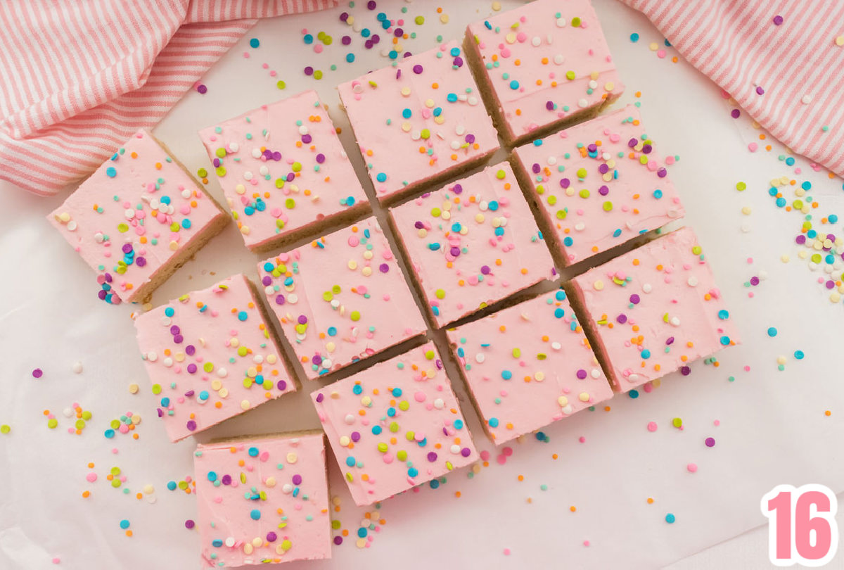 Overhead shot of 12 Sugar Cookie Bars sitting on a white table surrounded by sprinkles, and a pink striped kitchen towel.