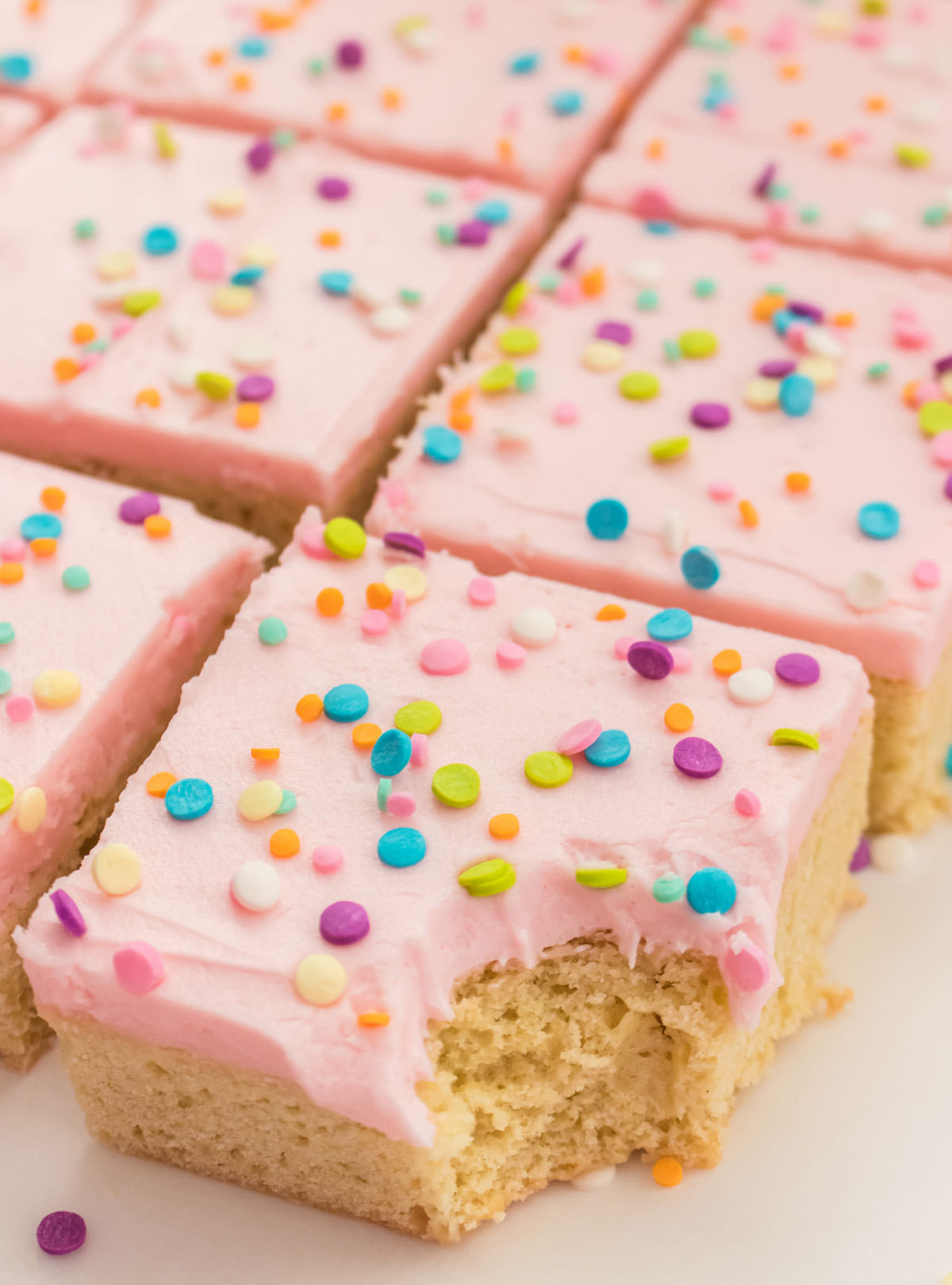 Closeup on rows of Sugar Cookie Bars covered in pink buttercream frosting and pastel sprinkles - the first one with a bite taken out of it.