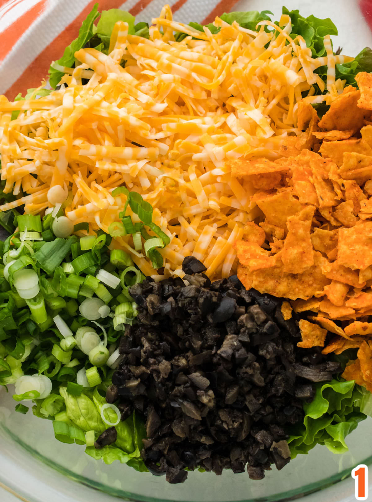Closeup on a glass bowl filled with the main ingredients of the Dorito Taco Salad including lettuce, grated cheese, Doritos, chopped olives and chopped green onions.