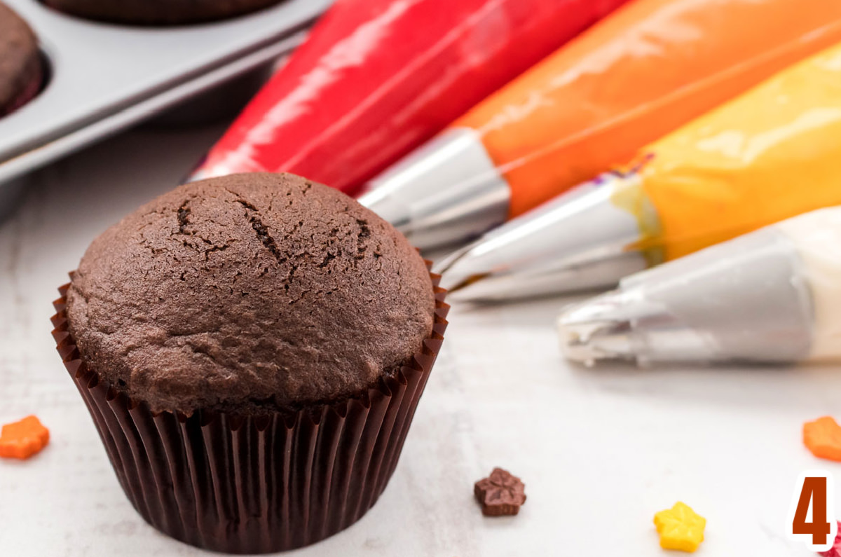 A single unfrosted cupcake sitting on a white table next to frosting decorating bags filled with red, orange, gold and white buttercream frosting.