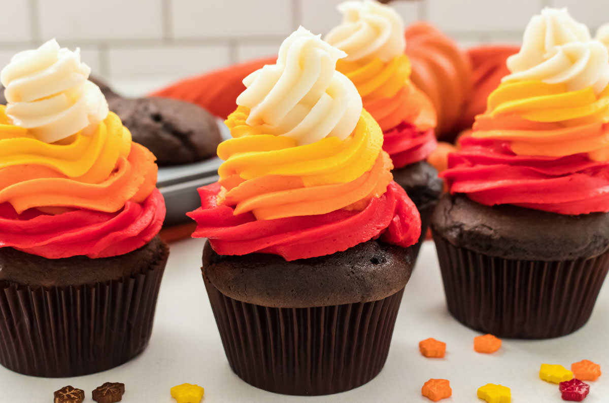 Four Harvest  Thanksgiving Cupcakes sitting on a white table in front of a pan of chocolate cupcakes.