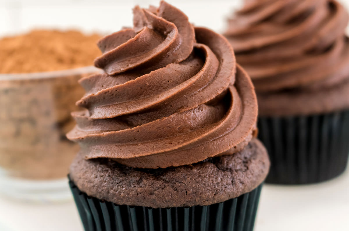 Closeup two cupcakes frosted with The Best Chocolate Buttercream Frosting sitting on a white table in front of a glass bowl filled with cocoa powder.