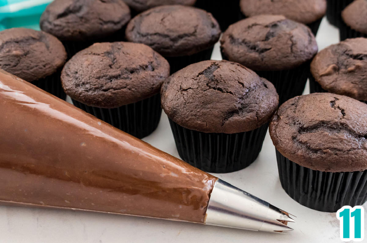 A frosting decorating bag filled with Chocolate Frosting sitting next to a batch of unfrosted chocolate cupcakes.