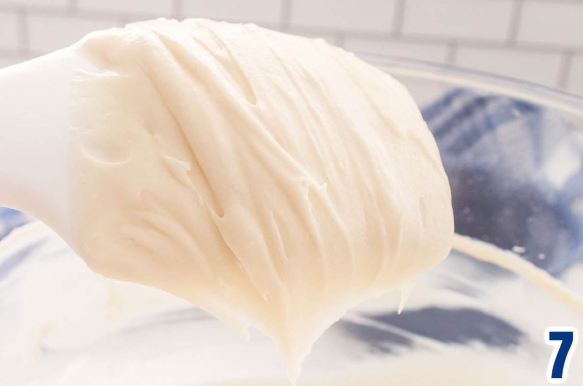 Close up on a white spatula covered with Cream Cheese Icing that is hovering above a glass mixing bowl that is sitting on a white table.