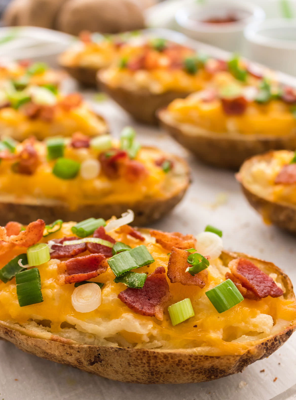 Closeup of eight Twice Baked Potatoes sitting on a cookie sheet surrounded by white ramekins filled with bacon and green onion.