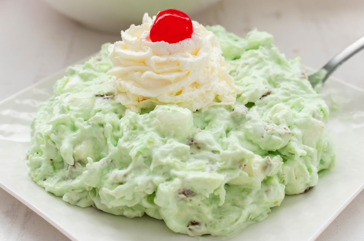 Closeup on a white plate filled with Watergate Salad topped with a dollop of whipped cream and a single Maraschino Cherry.
