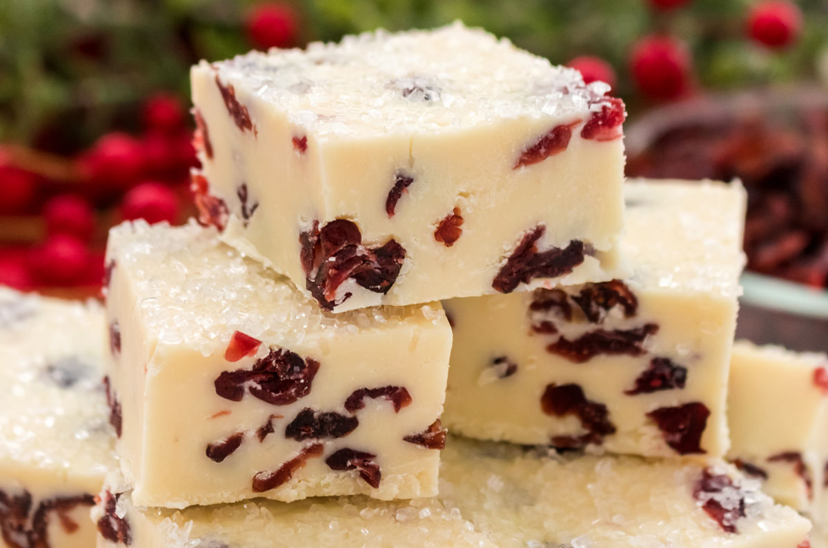 Closeup on three White Chocolate Cranberry Fudge pieces sitting on a stack of more fudge in front of cranberry decorations.